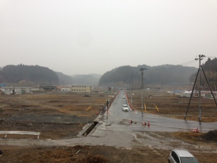 霧がかかった曇り空の下で山々に囲まれた広い荒地の中を一本の舗装道路がまっすぐに伸びており、その道路を数台の車が走っており、道路を挟んで右側に伊里前福幸商店街の建物が写っている写真