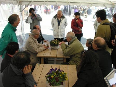 テントの中で、花が飾られた木製のテーブルを囲んで数人の大人が座り、周囲には他の大人たちが立って話し合いをしている様子の写真