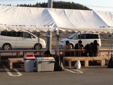 駐車場に設置されたテントの下にベンチとテーブルが設置されており、数人が座って話をしているコミュニティスペースの様子の写真