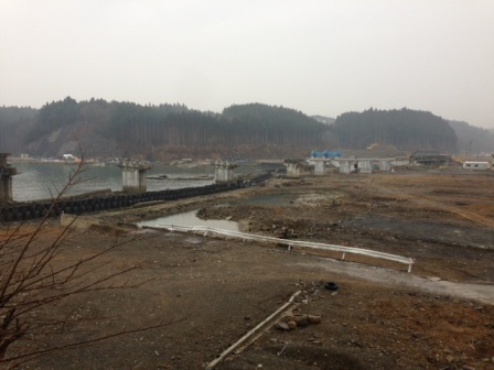 霧が立ちこめる空の下、川沿いに橋脚だけが立ち並び、その周囲には木々の茂る山々と重機や資材が点在する造成中の広い工事現場が広がっている様子の写真