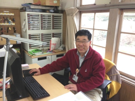 明るい窓辺の席で赤い上着を着た鈴木清美さんがパソコンの前に座って笑顔でカメラを向いている写真