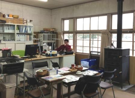 大きな窓から自然光が差し込む室内で、薪ストーブが窓側に設置され、奥では赤い上着を着た鈴木清美さんがデスクでパソコン作業をしている事務所内の写真