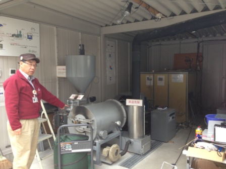 円筒形や円筒状の金属部品が組み合わさり、配管や煙突のような管が付いた発電装置の前に立った鈴木清美さんが指差して説明している写真