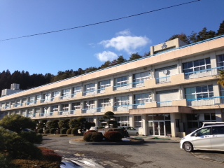 青空の下、三階建てでベランダのある校舎が横長に広がり、前庭には整えられた植木や数台の車が停まっている志津川高校の外観写真