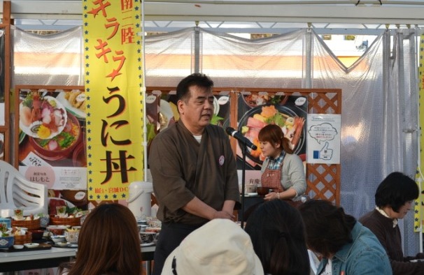 「キラキラうに丼」と書かれた黄色いのぼりの前に立った茶色い作務衣を着た高橋修さんががマイクの前で話しをしている写真