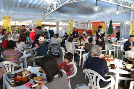 白いプラスチック椅子とテーブルが並ぶ広い飲食スペースでで多くの人が食事を楽しんでいる様子の写真