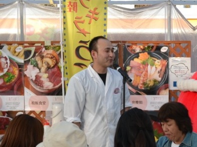 白い調理服を着た男性がマイクの前で話をしている写真