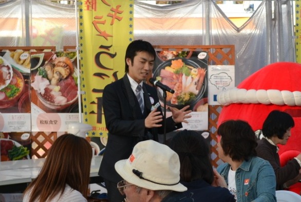 黒いスーツを着た男性がマイクの前に立ち、身振り手振りを入れて話をしている写真