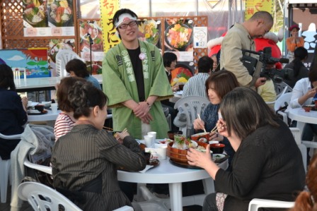 白い丸テーブルを囲んで4人の女性が食事をしているそばで、緑の法被を着た男性が笑顔で立ち話をしている写真