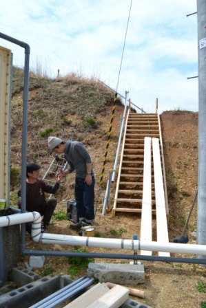 斜面に設置された木製の階段に2枚の細長い木の板が立てかけられており、その前で、二人の男性が相談しながら作業を行っている様子の写真