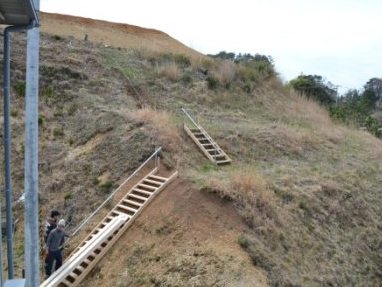 山の斜面に二か所の木製階段が設置され、下の階段のそばに二人の男性が作業を行っている様子が写っている写真
