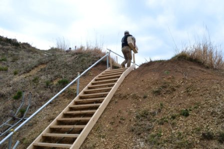 山の斜面に設置された手すり付きの木製階段を、荷物を持った男性が上っている様子の写真