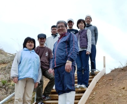 手すり付きの木製階段の上で、7人の男女が並んでカメラに向かって笑顔を見せている写真