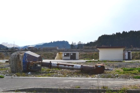 地面に倒れた細長い物があり、その奥に2件の建物が建っている広い空き地の写真