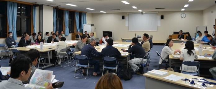 広い会議室で、多くの参加者が複数設置されたテーブルを囲んで座り、資料を見ながら活発に話し合っている様子の写真