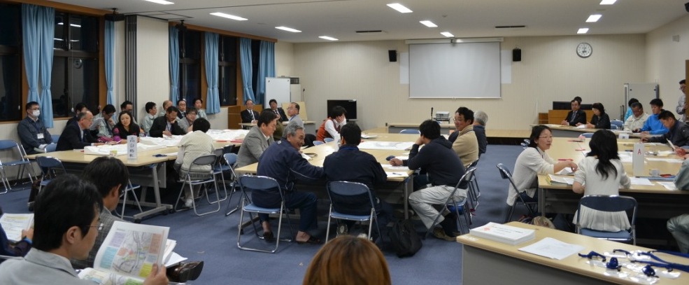 広い会議室で、多くの参加者が複数設置されたテーブルを囲んで座り、資料を見ながら活発に話し合っている様子の写真