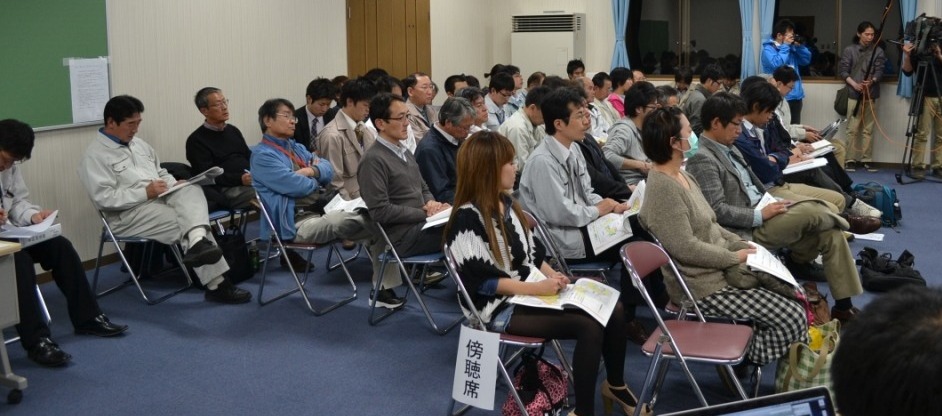 会場の後方に設けられた傍聴席に、多くの人々が座って話を聞いている様子の写真