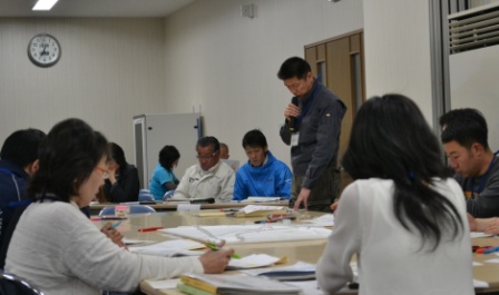 会議室で複数の人がテーブルを囲んで座っていて、一人の男性がマイクを持って立ち上がり話している様子の写真