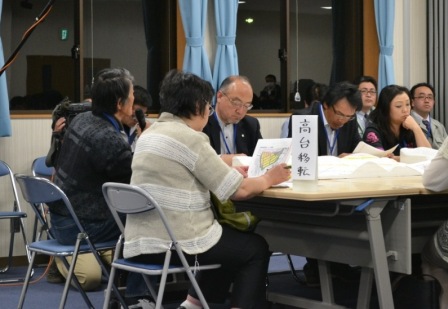 「高台移転」と書かれた紙が立てられたテーブルを囲んで数人が座っており、地図や資料を見ながら話し合っている様子の写真