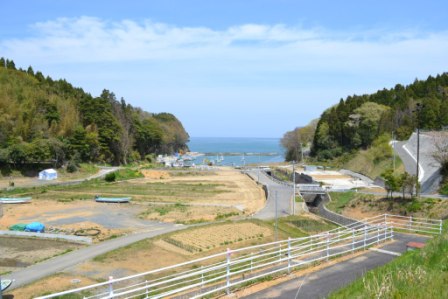 緩やかな坂道が海へと続き、空き地や畑の周囲は山々に囲まれている写真