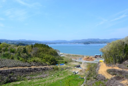 青い空の下で遠くに連なる山々を背景に、海が広がり手前には草地や木々が生い茂った風景を高い所から撮影した写真