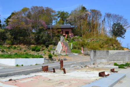 手前にはコンクリートで囲まれた空き地があり赤い手すりのついた階段を登った先に木々に囲まれた戸倉神社が見える風景写真