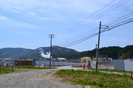 山々を背景に、中央に電柱と電線が立ち、白いフェンスで囲まれた部分にコンクリートなどの瓦礫が山となっており白い煙が見えている写真