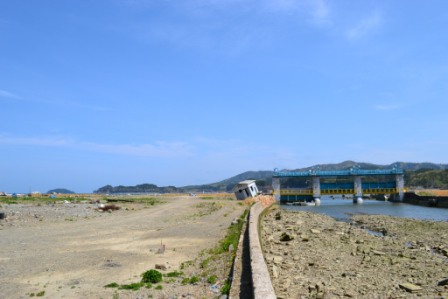 広い土地に右側に川とダムがあり堤防のそばに1件の斜めになった家がある写真