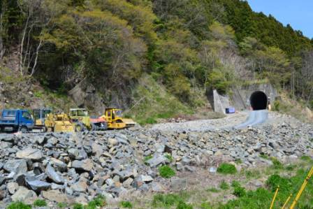 トンネルの入り口の前に複数の工事車両が並び、周囲には緑豊かな木々が広がり、地面には大きな石や岩が散らばっている写真