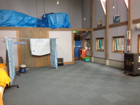 広いカーペット敷きの室内で、右端にはペレットストーブがあり、左には布団や毛布が置かれ壁際の棚の上にはブルーシートがかぶせられた荷物が載っている室内の写真