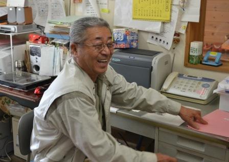 事務所の席に座って、笑顔を見せている菅原辰雄さんの写真