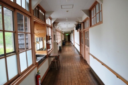 木枠の大きな窓が並ぶ明るい廊下が奥まで続いている校舎の廊下を写した写真