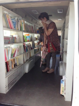 移動図書館の後方入口から撮影された車内には、左右の棚にたくさんの本が並べられ、二人の女性が本を手に取りながら本を探して見ている様子を写した写真