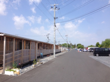 晴れた日の青空の下、左側に仮設住宅が連なり、右側には砂利の駐車スペースに車が並んでいる停車予定地を写した写真
