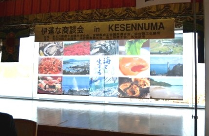 「伊達な商談会 in KESENNUMA」と書かれた横断幕の下に、海産物や自然風景、観光地の写真がコラージュ状に並べられたステージ背景が掲げられている様子を写したイベント紹介パネルの写真