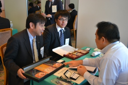 スーツ姿の男性2人が座りながら、対面のバイヤーに海産物のギフト商品を箱に入れて紹介している様子を写した商談中の写真