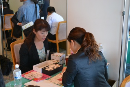 スーツ姿の女性がパッケージされた商品サンプルを机に並べて、向かいの女性に熱心に説明している様子を写した商談の様子の写真