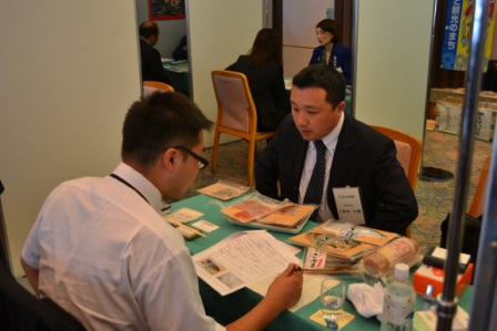 スーツ姿の男性が、水産加工品のパッケージを机に並べて書類を見ながら説明し、相手のバイヤーが真剣に話を聞いている様子を写した個別商談の写真