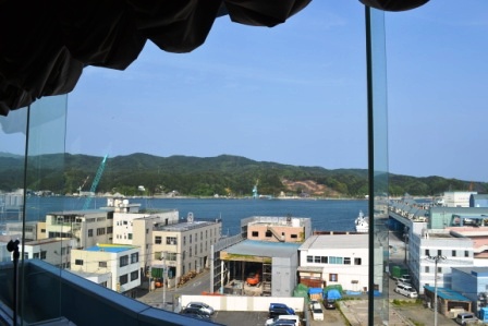 会場の大きな窓から見える外の景色として、海に面した気仙沼の港町の建物や停泊中の船、その向こうの山並みまでを一望できる様子を写した気仙沼港の風景の写真