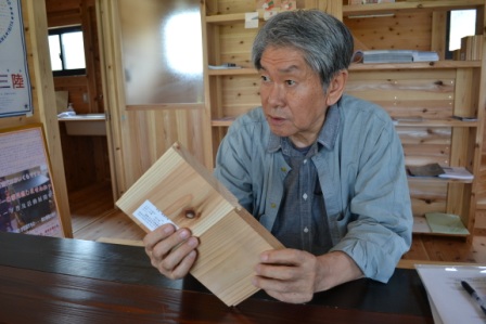 木の板材を手に持ち、その特徴について話している年配の藤村望洋さんが、木材に囲まれた室内で説明している様子の写真