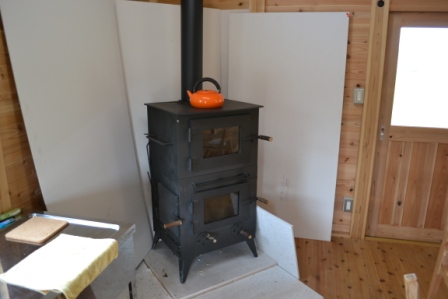 室内の角に黒い薪ストーブが設置されており、上にはオレンジ色のやかんが載っている、暖房と調理を兼ねたストーブの写真