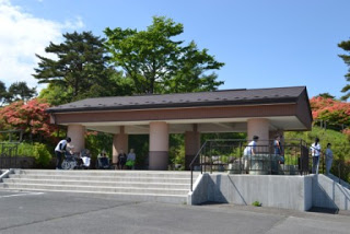 茶色い屋根と薄いピンク色の柱のある東屋風の休憩施設の周りに人々が集まっており、ツツジと木々に囲まれた中で憩う様子を写した休憩所の写真