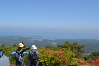 展望所でリュックと帽子を身につけた二人の人物が説明板の前に立ち、海や島々の景色を指さして眺めている様子を写した展望台の写真