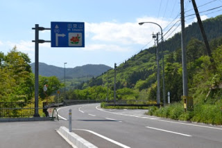 山あいの道路に設置された青い案内標識に「母王山」と書かれており、周囲には電柱と緑の木々が立ち並ぶ中、カーブを描く道路が奥へと続いている様子を写した道路標識の写真