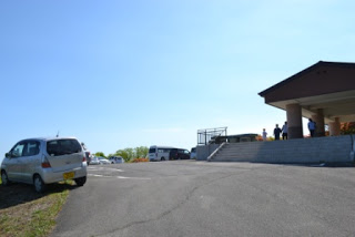 青空の下、右側に屋根のある施設があり、左には駐車された車や観光バスが見える広い舗装の駐車場に数人の人が集まっている様子を写した駐車場の写真