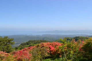 赤やオレンジのツツジが咲く丘の上から、遠くの海や島々を一望できる景色が広がり、空と海の青が溶け合うように見える様子を写した展望の写真