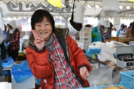 オレンジ色のジャンパーを着た女性が笑顔でピースサインをして写っている写真