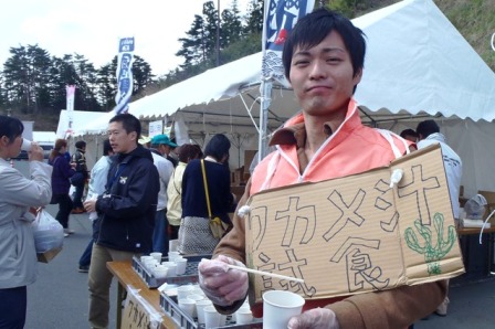 屋外のイベント会場で、ピンクのジャケットを着た男性が「ワカメ汁試食」と書いた段ポールの看板を持ち、試食用のカップを手にしている写真