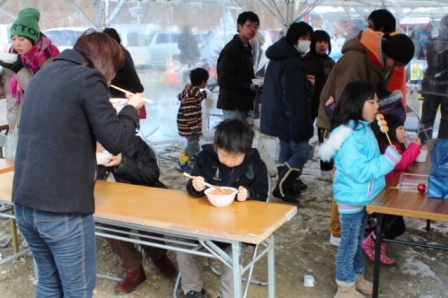 冬の屋外イベント会場で来場者の家族や子どもたちがテント下に設置された机の前で温かい食べ物を食べたり、団子を食べたり、飲食を楽しんでいる様子の写真