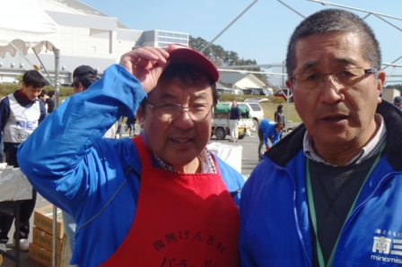 屋外の会場で青いジャンパーを着た二人の中年男性がカメラに向かって笑顔を見せて写っている写真